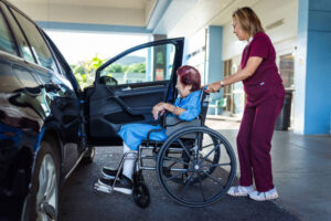 A senior in a wheelchair being assisted outside a Houston hospital or clinic (e.g., Houston Methodist or Memorial Hermann).
