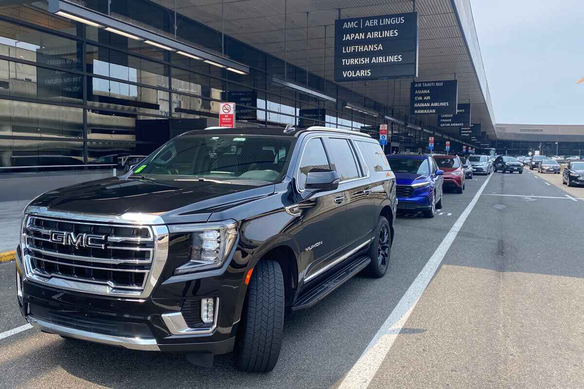 Black luxury sedan parked at airport curb, ready to pick up arriving passenger for intercity car service.